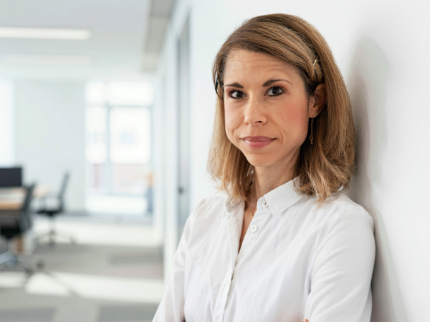 Frau mit langen, blonden Haaren in einem modernen Büro, lächelnd und nachdenklich.